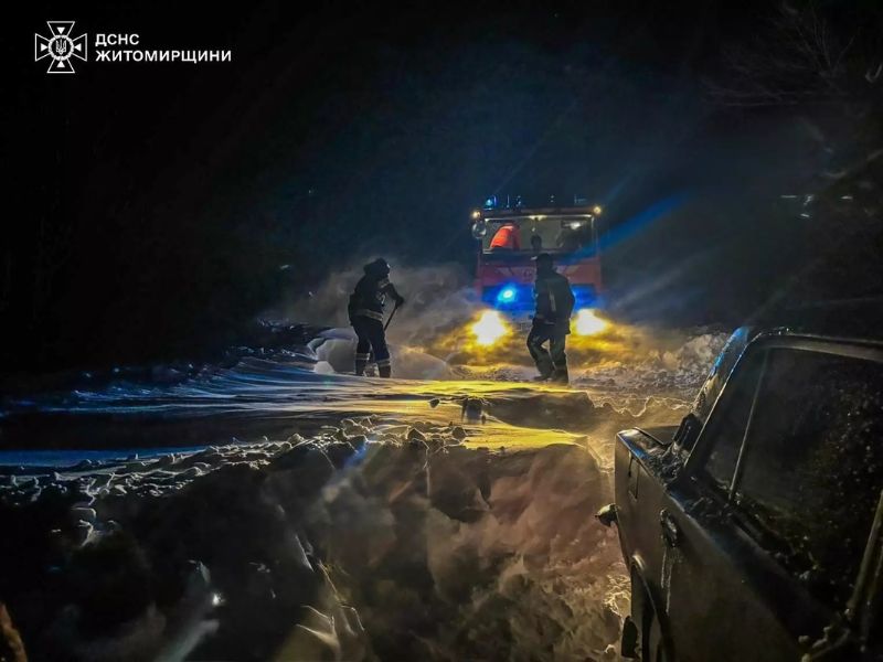 Спасатели ГСЧС в Житомирской области ночью расчищают снежные заносы перед застрявшей легковой машиной. Видны яркие фары спецтехники и работа спасателей в зимней экипировке.