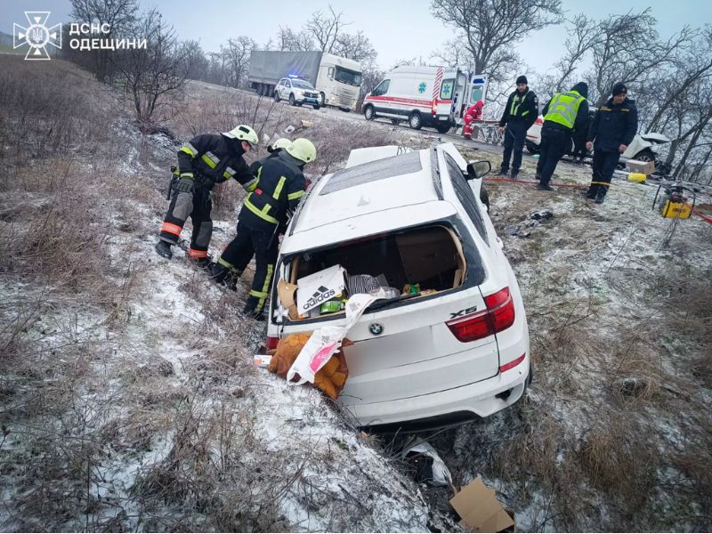 Белый автомобиль BMW X5 после аварии в кювете на заснеженной обочине, спасатели ГСЧС Одесской области работают на месте происшествия, скорая помощь и полиция на трассе.