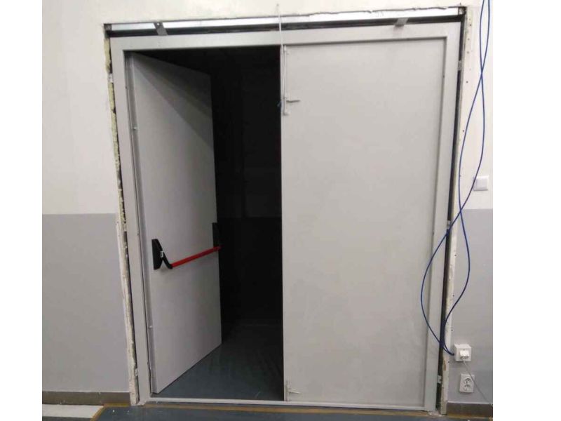 В білу стіну вмовтовані сірі двері із системою антипаніка із червоною дверною ручкою