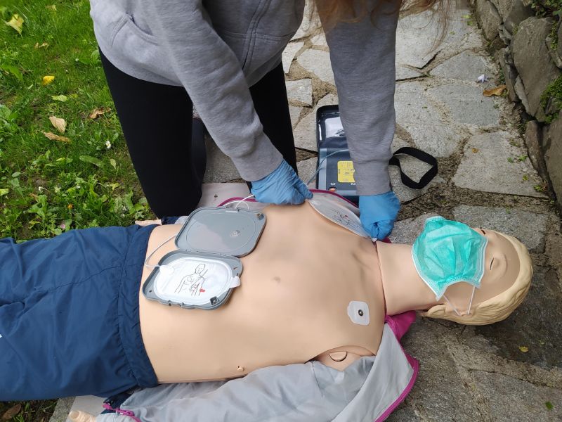 Людина в сірому худі прикріплює частини дифібрилятора до медичного манікену у якого відсутня одежа на верхній частині тіла, але в цього манікена є сині штани.