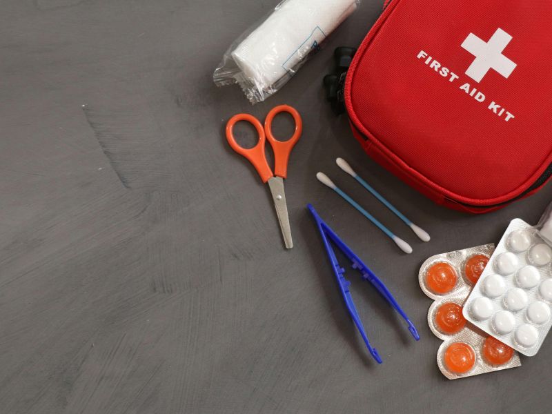На столі сірого кольору знаходяться різні медикаменти, ножиці, пінцет, вушні палички, бинти, таблетки білого та поморанчевого коьлору і відкрита сумочка аптечки червоного кольору.