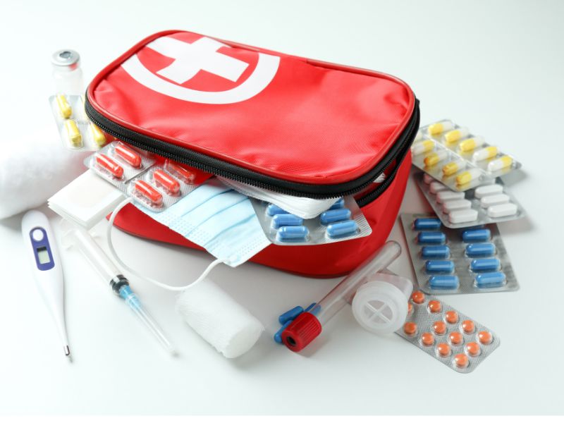 На білому фоні в центрі кадра знаходиться кейс червоного кольору із білим хрестом на ньому. Поряд із кейсом та у кейсі знаходяться різного роду лікумальні засоби як таблетки, шприци, бинт та пластир.