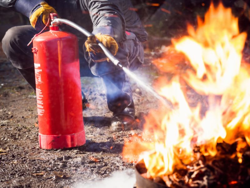 На корточках сидить чоловік у пожежному спорядженні та намагається загасити полум'я направляючи струмінь із вогнегасника із шлангом на джерело полум'я