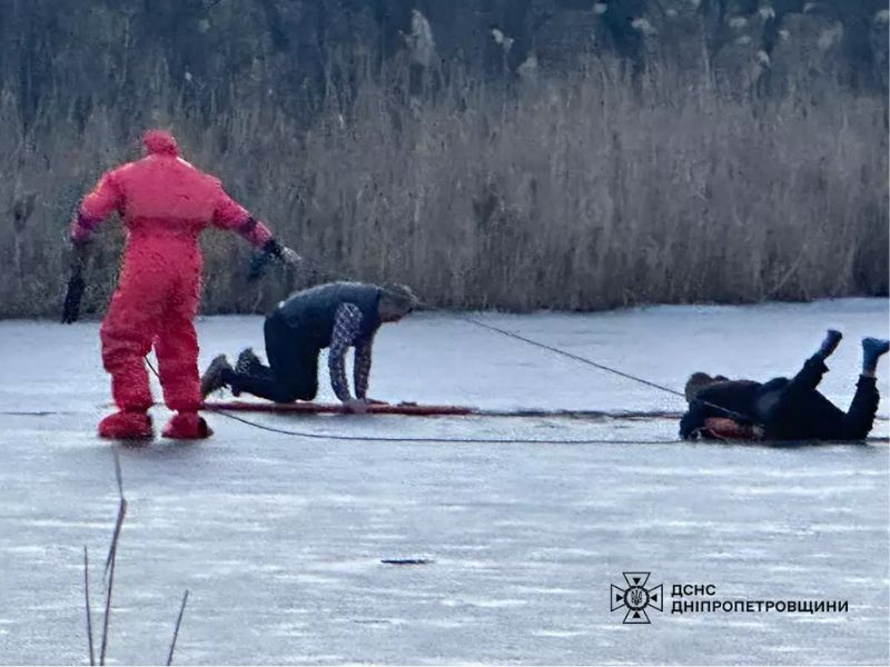 Спасатель ГСЧС в красном защитном костюме помогает двум мужчинам выбраться с тонкого льда. Люди ползут по замерзшему озеру, удерживаясь за веревки. Логотип ГСЧС Днепропетровской области.