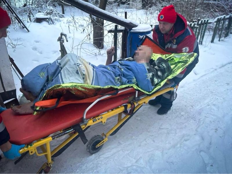 Медицинские работники эвакуируют мужчину на носилках по снегу. Пострадавший накрыт термоодеялом и синим пледом. Спасатель в красной форме и шапке сопровождает каталку в зимнее время.