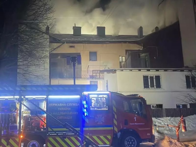 Пожежна машина ДСНС Івано-Франківська під час ліквідації загоряння у будинку. Нічна сцена з густим димом над дахом будівлі та спецтехнікою рятувальників на засніженій вулиці.