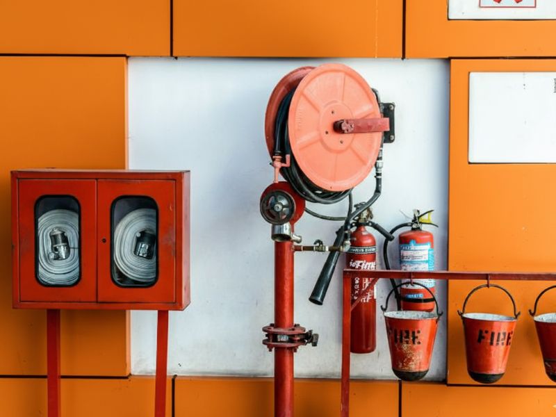 На фоні помаранчевої стінки находиться пожежний рукав, пожежні конусні відра, лопата і два вогнегасника