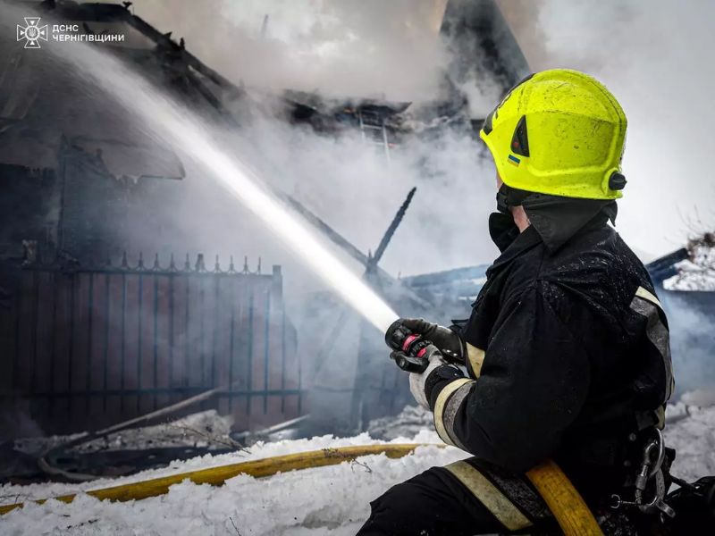 Пожарный ГСЧС в Черниговской области тушит сильный пожар зимой. Спасатель в защитной форме и ярком шлеме направляет струю воды из брандспойта на дымящиеся руины здания.