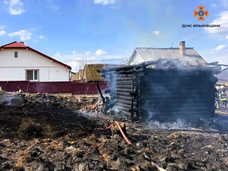 Обгоревшая деревянная постройка после пожара в Ивано-Франковской области. На месте работают спасатели ГСЧС, на земле видны пожарные рукава и выжженная трава.
