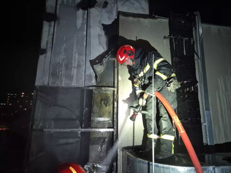Пожарный в защитной экипировке и красном шлеме тушит обгоревшие элементы здания из брандспойта в ночное время.