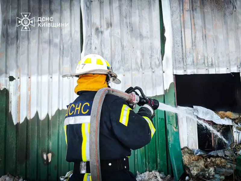 Пожарный ГСЧС Киевской области в защитном снаряжении тушит возгорание в металлическом ангаре, направляя струю воды из пожарного рукава внутрь помещения.