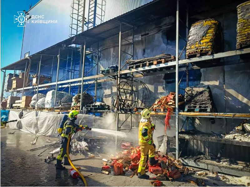 Два пожарных ГСЧС в защитной экипировке тушат огонь на складе под Киевом: один спасатель направляет струю воды на горящие остатки груза возле стеллажей.