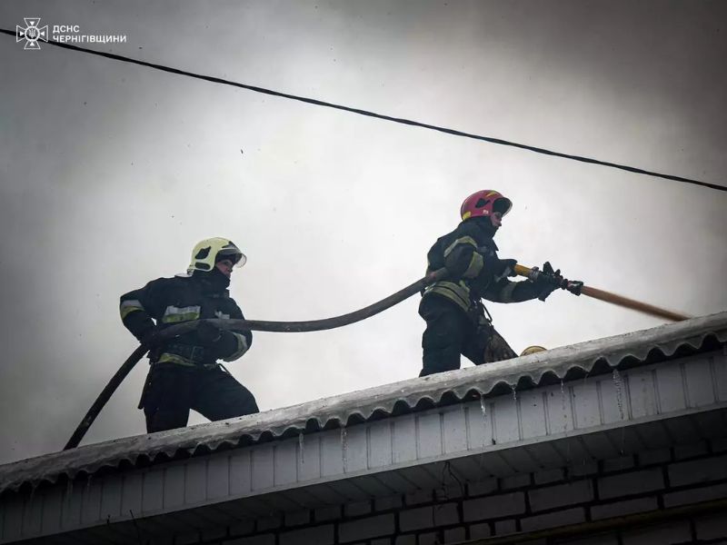 Рятувальники ДСНС Чернігівщини ліквідовують пожежу на даху будинку. Двоє вогнеборців у спецспорядженні та касках працюють з пожежним рукавом на засніженій покрівлі, подаючи воду на вогонь.