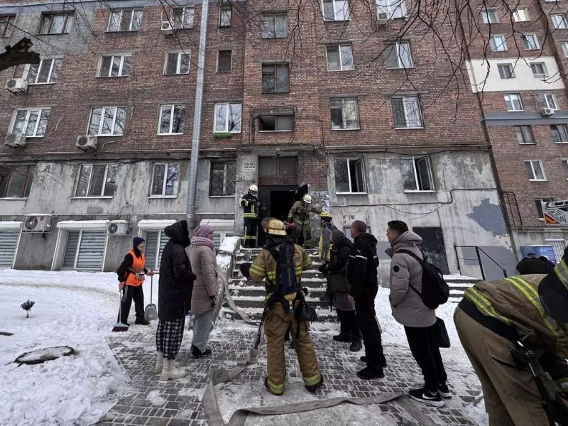 Рятувальники ДСНС працюють на місці пожежі у житловому багатоповерховому будинку взимку. Вогнеборці у захисному спорядженні біля під'їзду, розгорнуті лінії пожежних рукавів на снігу.