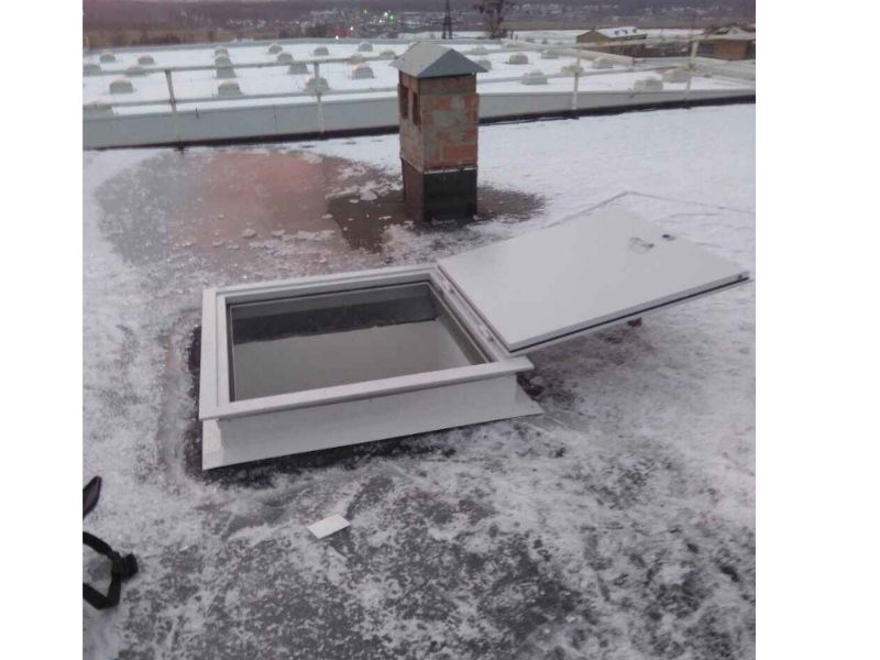 На криші будівлі встановлено пожежний люк білого кольору, що є відкритим. Навколо видніється вечірнє місто та сніг із льодом.