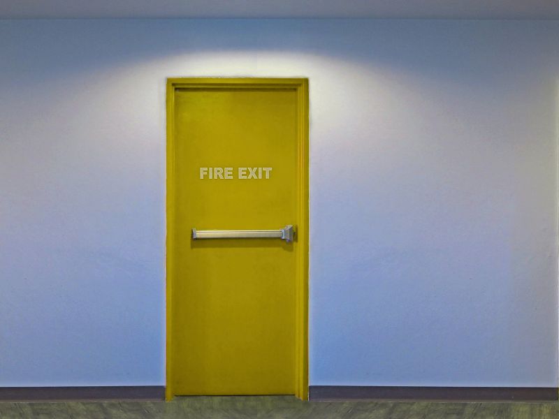 На фоні білої стіни жовтого кольору пожежна дверь із системою антипаніка
