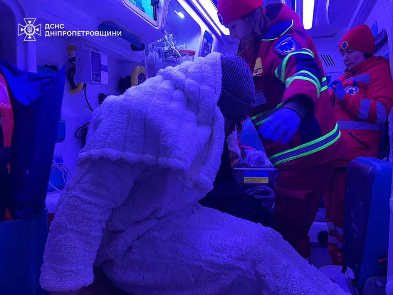 Внутри спасательного автомобиля ГСЧС медики оказывают помощь человеку, завернутому в теплый халат. Синее освещение в салоне спецтранспорта.