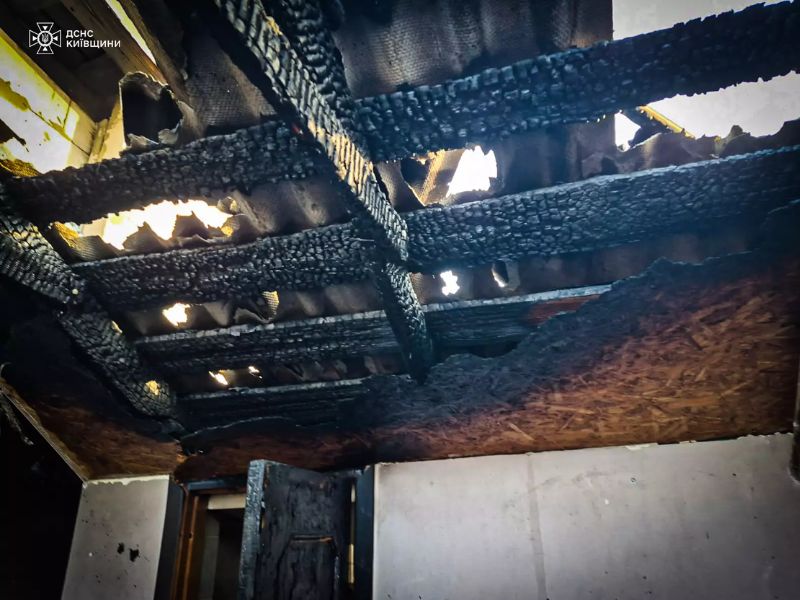 Внутренний вид обгоревшей крыши дома после пожара: обугленные до черного цвета деревянные балки, прогоревшие отверстия в кровле и копоть на стенах.