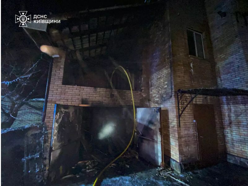 Вид на сгоревший кирпичный гараж и второй этаж частного дома после пожара ночью, видны обгоревшие балки крыши.