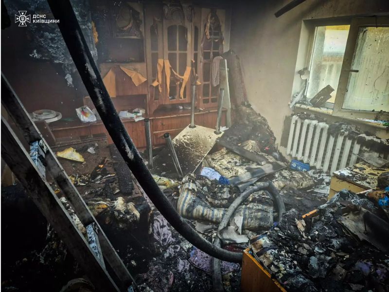 Последствия пожара в жилой комнате в Киевской области: обгоревшая мебель, черные от копоти стены и пожарный рукав на переднем плане.