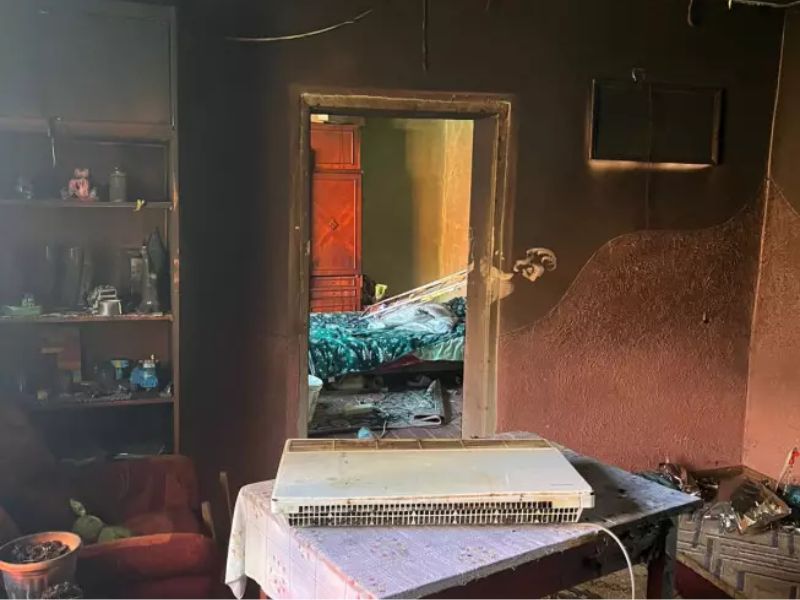 Интерьер комнаты после пожара: стены в черной копоти, на столе лежит электрический обогреватель, послуживший причиной возгорания. Вид через дверной проем на обгоревшую спальню.