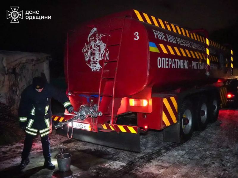 Пожарный ГСЧС наполняет металлическое ведро водой из крана большой красной автоцистерны с надписью Fire and Rescue Service of Ukraine. Ночь, снег на дорогах.