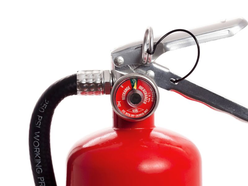 На білому фоні дуже наближено ми бачимо верхню частину вогнегасника із трубкою та механізмом спуску заряду і також манометр.