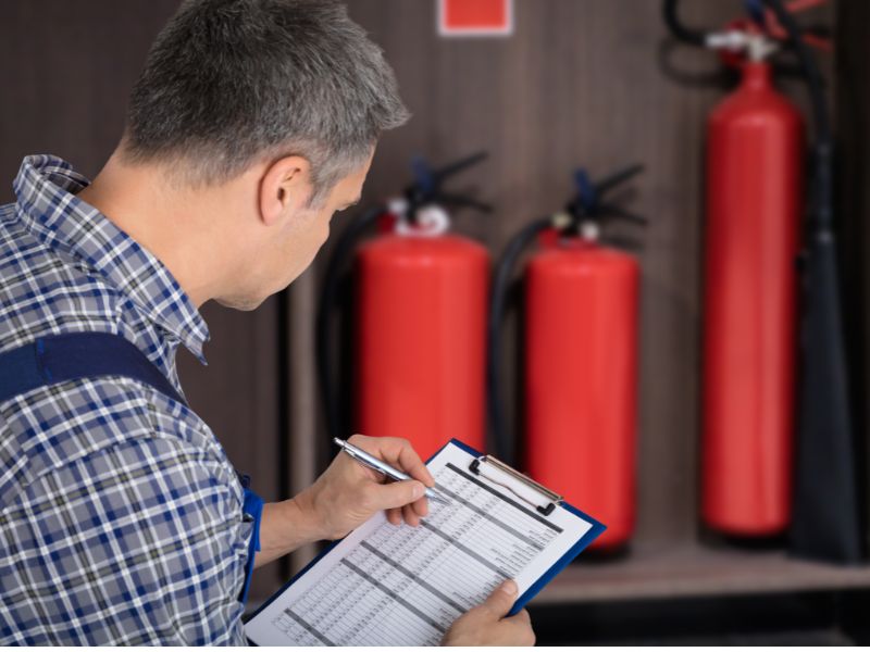 Чоловік середнього віку в клітчастій сорочці чидить в пів оберта. Він має багато сивого волосся та коротку стрижку. Вйого руках планшет із паперами, які він заповнює. На фоні знаходиться стіна коричневого кольору на якій закріплені три вогнегасники і видно знак "вогнегасник"