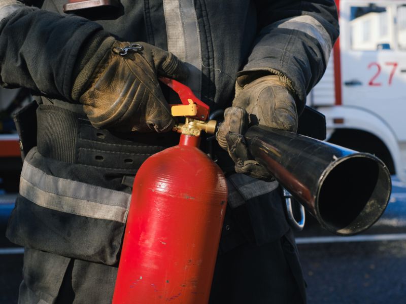 Ми бачимо середню частину тіла чоловіка молодога вдягненого в пожерний одяг, що тримає в руках вуглекислотний вогнегасник. В нього на руках вдягнуті рукавички. На задньому фоні можна бачити частково вулицю та припарковану карету швидкої допомоги. 