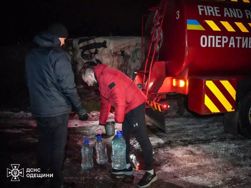 Местные жители набирают воду в большие пластиковые бутыли и ведра из красной автоцистерны ГСЧС Одесской области ночью на заснеженной улице.