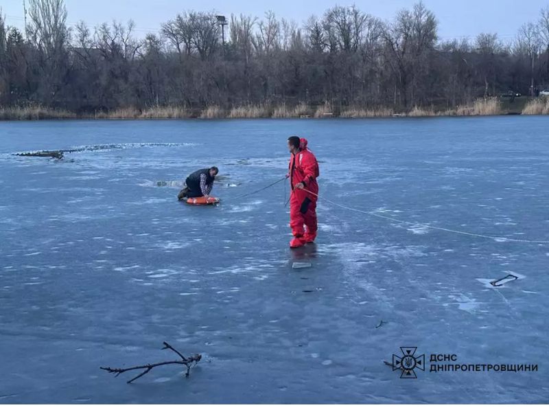Спасатель ГСЧС в красном защитном костюме на льду помогает человеку, который держится за спасательный круг в полынье. Процесс спасения на тонком льду в Днепропетровской области.