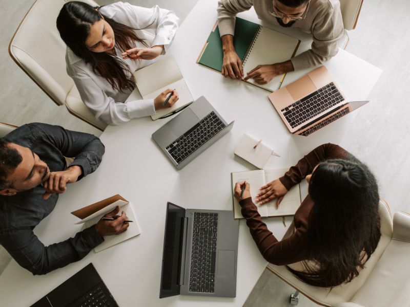 За столом сидять чотири людини із відкритими ноутбуками сірих кольорів, дві жінки та два чоловіка, вони всі знаходяться у офісі