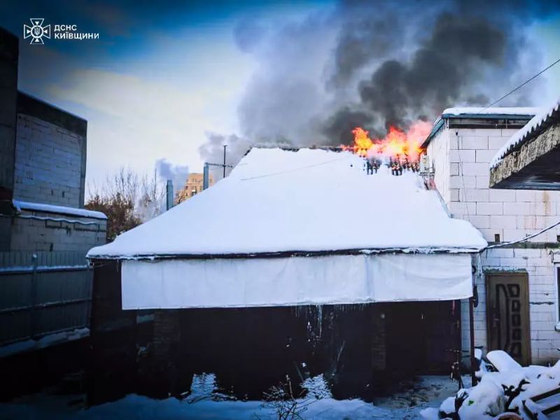 Пожар на крыше частного дома зимой в Киевской области: из-под заснеженной кровли вырывается открытое пламя и густой черный дым.