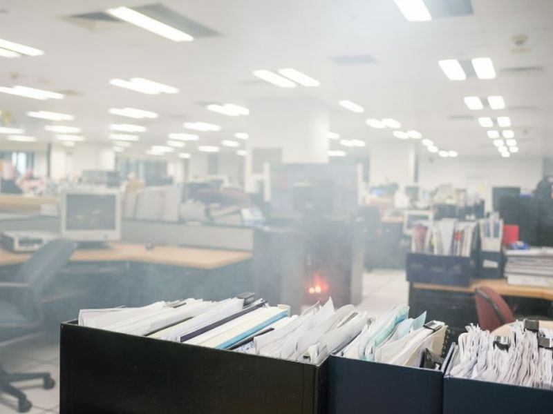 На передньому плані папки та докуенти на фоні офісного антуражу, також на задньому плані видно задимлення та початок пожежі