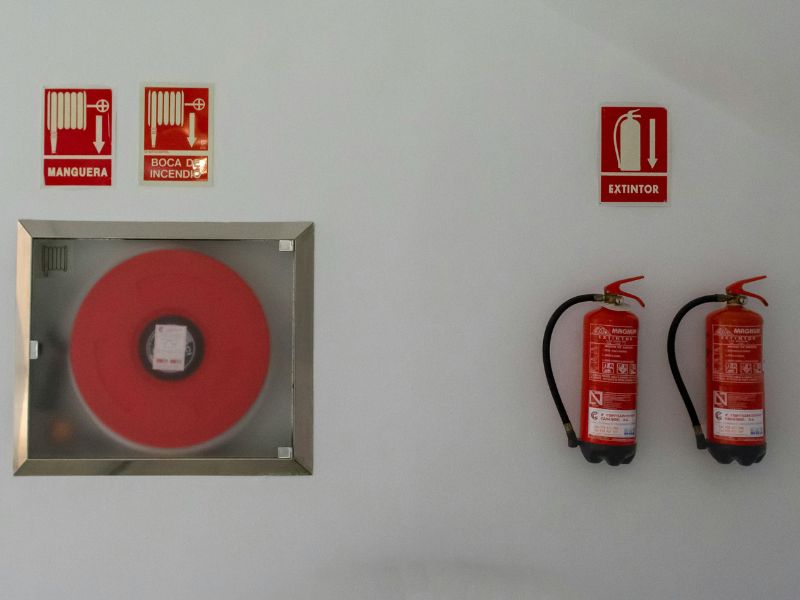 На фоні бфлої стіни знакодиться пожежна шафа із пожежним рукавом всередині, на стіні є позначки пожежного гідранту і також у лівій частині кадру знаходяться два вогнегасники червоного кольору