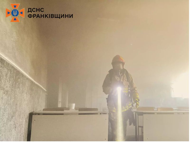 Пожарный в полном защитном снаряжении с фонарем в руках идет через густой дым в задымленном помещении класса.