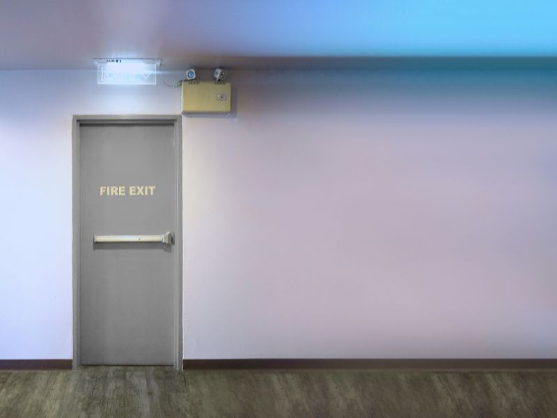 На фоні білої стіни протипожежні двері із ручкою антипаніка над ними позначка евакуаційного виходу та камера спостереження