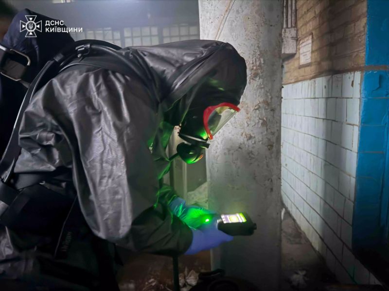 Сотрудник ГСЧС в полном костюме химической защиты и противогазе использует электронный газоанализатор для проверки уровня загрязнения в подвальном помещении.