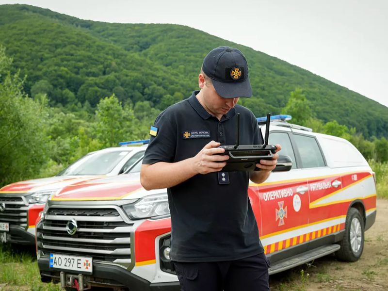 Співробітник ДСНС Закарпаття використовує пульт управління безпілотником для аеророзвідки території на тлі оперативно-рятувальних автомобілів.