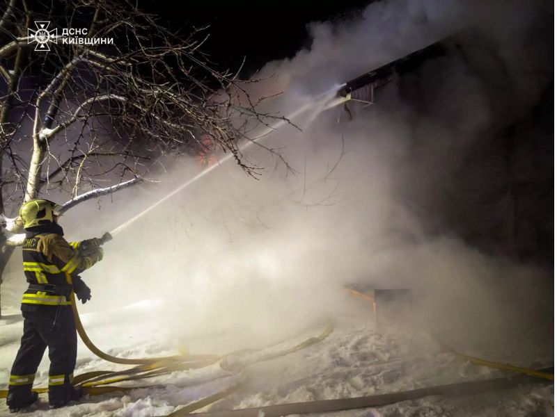 Пожарный ГСЧС Киевской области направляет струю воды из брандспойта на охваченное дымом здание ночью зимой.