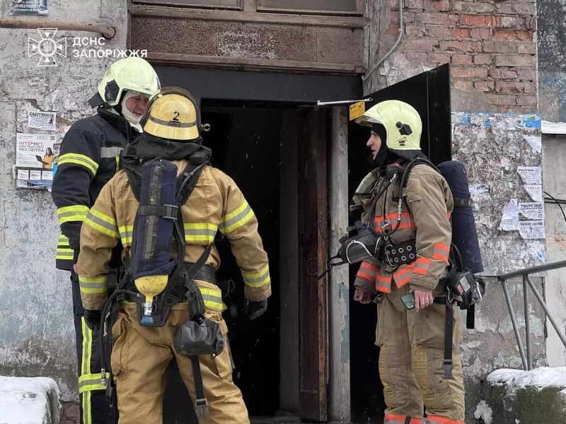 Троє пожежників ДСНС Запоріжжя у захисному екіпіруванні та дихальних апаратах обговорюють дії біля входу до під'їзду житлового будинку взимку.