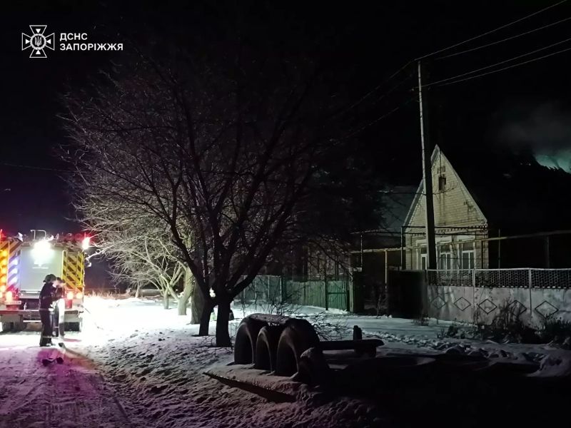 Пожежний автомобіль ДСНС з увімкненим освітленням на засніженій вулиці вночі. На задньому плані приватний будинок, де триває ліквідація пожежі підрозділами Запоріжжя.