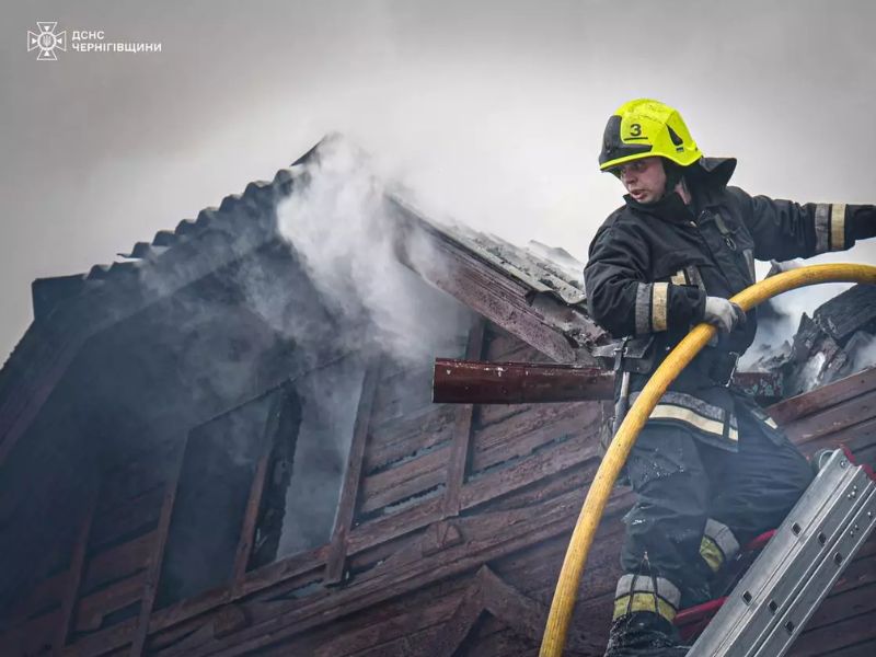 Рятувальник на драбині здійснює гасіння палаючого даху дерев'яного будинку. Крупний план співробітника ДСНС Чернігівщини у захисному шоломі з пожежним рукавом під час ліквідації задимлення на висоті.