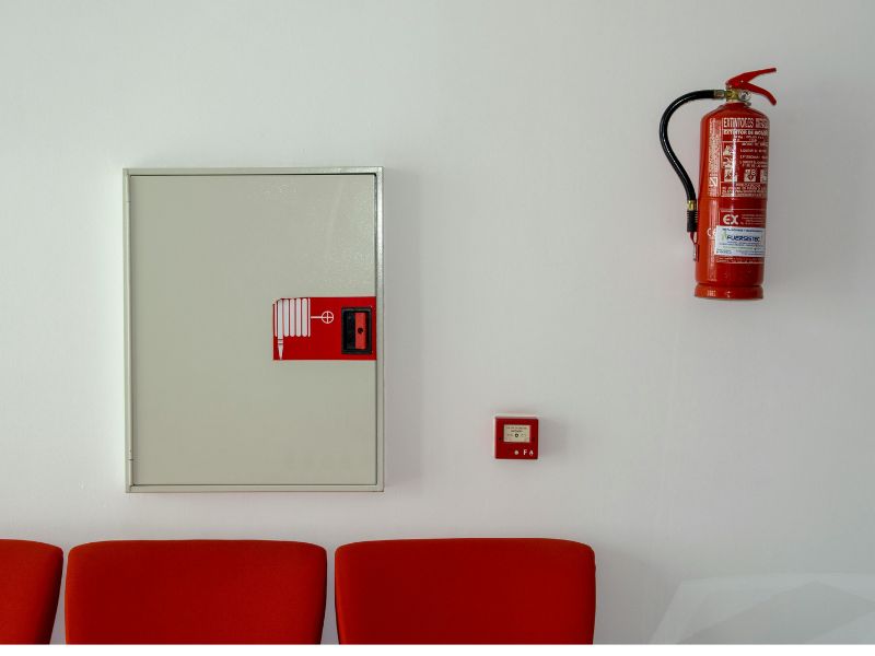 На фоні білої стіни знаходиться два червоних стільця, пожежна сигналізація, та червоний вогнегасник закріплений на стіну.