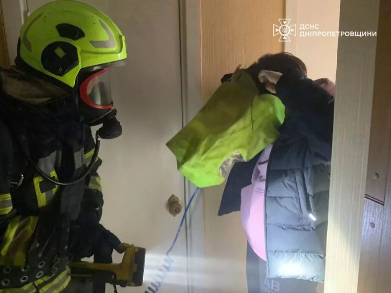 Пожарный ГСЧС надевает на женщину спасательный капюшон для защиты органов дыхания во время эвакуации из задымленного помещения, спасатель в защитном шлеме и дыхательном аппарате.