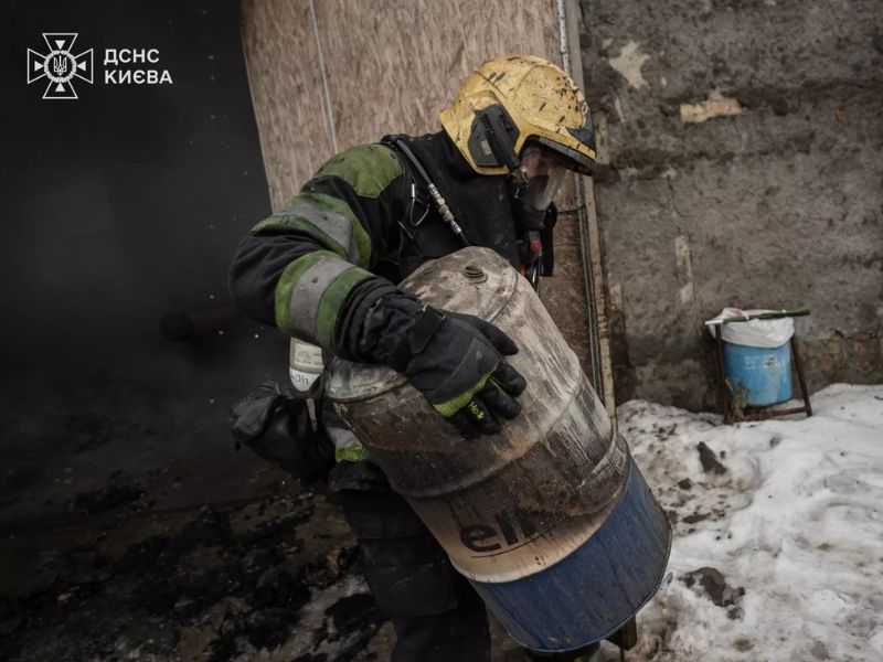 Рятувальник ДСНС Києва у повному бойовому спорядженні та дихальному апараті виносить обгорілу металеву бочку з місця пожежі на фоні снігу та згарища.