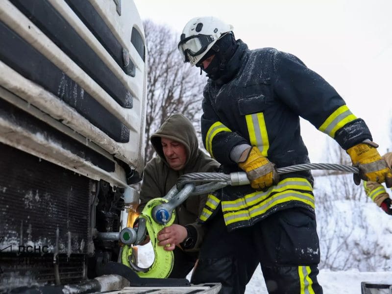 Крупный план: спасатель ГСЧС в защитных перчатках и водитель крепят тяжелый стальной трос с ярко-желтым крюком к передней части грузового автомобиля в снегу