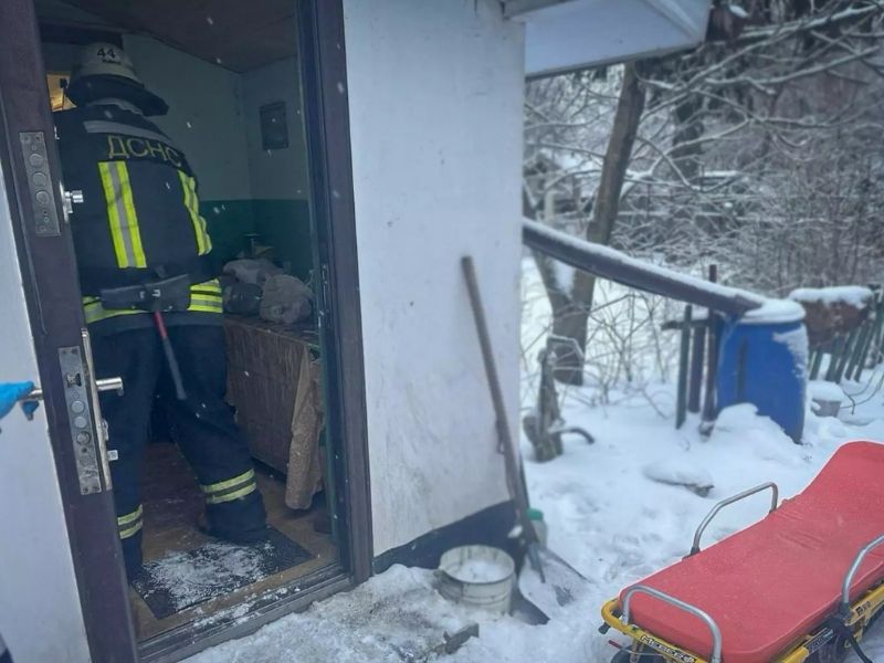 Пожарный ГСЧС в защитной экипировке заходит в дом в зимнее время. На переднем плане стоят пустые медицинские носилки на снегу, готовые к эвакуации пострадавшего.