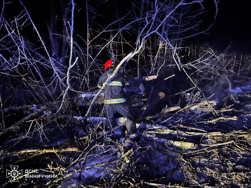 Спасатель ГСЧС в светоотражающей форме и красном шлеме расчищает завалы из поваленных деревьев и веток ночью в Винницкой области.