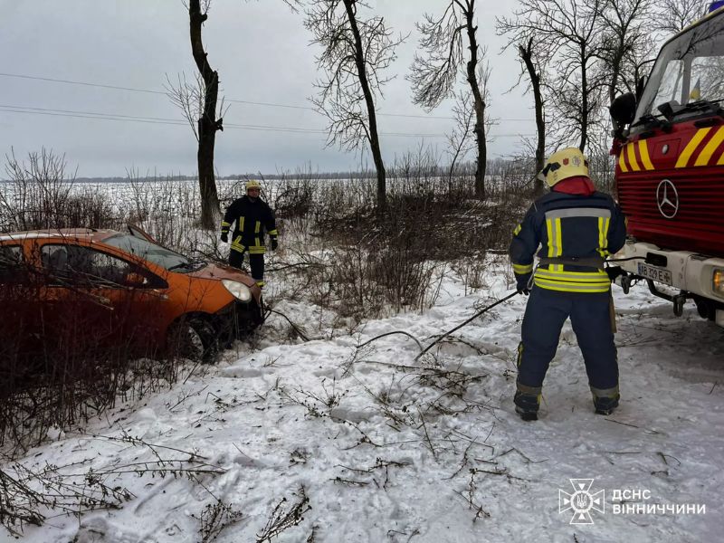 Пожарные ГСЧС Винницкой области вытаскивают оранжевый легковой автомобиль из заснеженного кювета после аварии. Спасатели используют спецтехнику в зимних условиях.